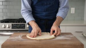 A person shapes and stretches pizza dough by hand on a floured wooden surface