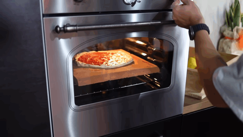 A person places a pizza in the oven on a baking stone