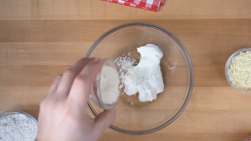 Someone adding a small amount of flour to Greek yogurt for pizza dough preparation