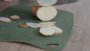 A knife slices a potato into thin rounds on a green cutting board