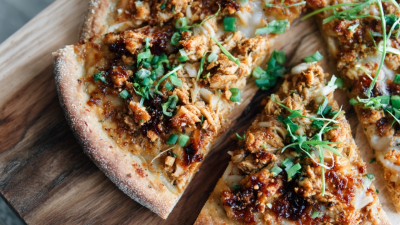Pizza topped with tofu crumbles, jackfruit pieces, fresh greens, and a drizzle of savory sauce on a crisp crust